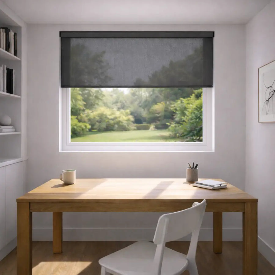 Modern home office with a Charcoal Sunscreen Roller Shade partially open, showing a bright sunbeam on a wooden desk and a view of a green garden.